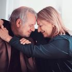 Ein älteres Paar umarmt sich herzlich, beide lächeln mit geschlossenen Augen und berühren liebevoll die Köpfe.