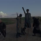 Vier Personen auf einem Feldweg, eine Puppe auf Stangen, Abendstimmung mit Wolken und Telegrafenmasten.