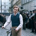 Ein junger Mann in altmodischer Kleidung steht mit einem Fahrrad auf einem gepflasterten, belebten Hinterhof.