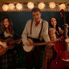 Drei lachende Personen unter Lichterketten spielen Gitarre, Banjo und Kontrabass in einem rustikalen, stimmungsvollen Ambiente.