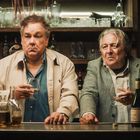 Zwei Männer stehen an einer Theke, halten Gläser. Getränke und Flaschen auf der Bar, Regal mit Gläsern und Flaschen im Hintergrund.