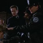 Ein Polizist hält eine Waffe; ein anderer drückt einen Mann gegen ein Auto. Nacht, städtische Umgebung, Blaulicht sichtbar.