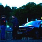 Frau mit "Police" Armband spricht mit Mann vor schwarzem Mercedes. Hintergrund: Bäume, Laternen, Zäune.