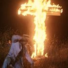 Ein brennendes Kreuz in der Nacht. Eine Person ist in Panik, umgeben von Flammen, auf einem Feld.