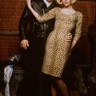 Ein Mann in schwarzer Lederjacke und Frau im Leopardenkleid vor einer Backsteinwand, eng umarmt.
