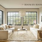 "ROOM BY ROOM" steht in einem stilvollen Wohnzimmer mit beigen Sofas, großem Fenster und Pflanzen. Teppich in der Mitte.