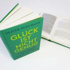 Auf einem grünem Buchcover steht: "OLIVER HOFFMANN, GLÜCK IST NICHT GENUG". Ein Buch liegt daneben offen.