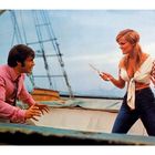 Zwei Personen auf einem Boot. Eine Frau mit Messer, trägt offene Bluse. Ein Mann in rosa Hemd wirkt überrascht.