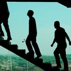 Drei Silhouetten von Menschen gehen eine Treppe hoch, vor einem hellen, blauen Himmel.