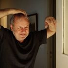 Ein älterer Mann mit grauem Haar lächelt. Er trägt ein dunkles T-Shirt und stützt sich an einer Tür ab.