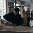 Ein Mann arbeitet konzentriert mit Werkzeugen in einer Werkstatt mit großen Fenstern und diversen Möbeln im Hintergrund.