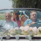Zwei Männer fahren in einem Cabrio, mit Blumen geschmückt. Zwei Frauen sitzen hinten. Alle schauen entspannt.