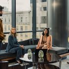 Vier Personen in einem modernen Büro mit großen Fenstern; zwei sitzen, zwei stehen, alle in formeller Kleidung.