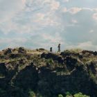 Keine Texte vorhanden. Eine Klippe unter blauem Himmel mit Wolken. Zwei Personen gehen entlang des Grats, umgeben von Natur.