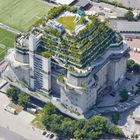 Ein großes, terrassenförmiges Gebäude mit viel Begrünung oben, neben einem Fußballplatz. Parkplatz und Straßen umgeben es.