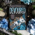 Ein Buchcover mit dem Titel "Devoured" von Jordan Stephanie Gray, verziert mit einer Krone, blauen Blumen und Ornamenten.