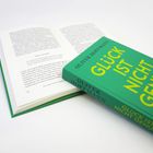 Das Buch "GLÜCK IST NICHT GENUG" von Oliver Hoffmann liegt auf einem offenen Buch.