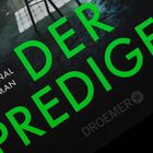 „Kriminal Roman„ „DER PREDIGER“ in grünen Großbuchstaben, „DROEMER“ unten. Hintergrund mit Licht und Schatten.