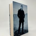 Ich kann den Text nicht erkennen. Es ist ein Foto eines Mannes an einem Ufer, das auf dem Einband eines Buches gedruckt ist.