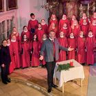 Ein Chor von Kindern in roten Gewändern, dirigiert von einem lächelnden Mann, steht auf einer Bühne mit einem Adventskranz.