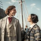 Zwei Personen in historischer Kleidung lächeln sich auf einem Pier an. Im Hintergrund ein Segelschiff und Zuschauer.