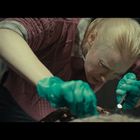 Eine Frau mit blonden Haaren und grünen Handschuhen untersucht konzentriert etwas mit einer Lampe.
