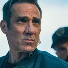 Zwei Männer im Freien, der vordere mit Nachdenklichkeit in Lederjacke, der hintere trägt eine Cap mit einem unscharfen Logo.