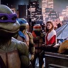 Vier humanoide Schildkröten mit bunten Masken stehen auf einem Dach vor einer Stadt, neben einer Frau und einer Ratte.