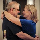 Eine lachende Frau und ein Mann umarmen sich; im Vordergrund eine Sektflasche.