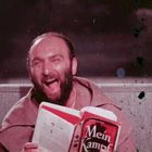 Ein Mann lacht laut, während er ein Buch mit dem Titel "Mein Kampf" in den Händen hält.