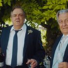 Zwei ältere Männer in eleganten Anzügen mit Blumenansteckern unter einem grünen Laubdach, einer hält eine Flasche.