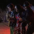 Ein blutverschmiertes Trio: eine Frau in einem roten Blumenkleid wird von zwei Männern gestützt, es ist Nacht mit Schnee.