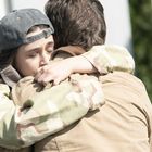 Zwei Personen umarmen sich herzlich, eine trägt eine Kappe und militärähnliche Jacke, die andere einen beigen Mantel.