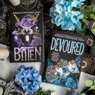 Links steht "BITTEN" mit einem Wolfsbild und lila Blumen, rechts "DEVOURED" mit einer Krone und blauen Rosen.