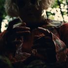 Eine Person mit blonder Perücke in einem Wald, trägt rote Kleidung mit blutähnlichen Spuren auf Händen.