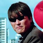 Ein Mann mit Sonnenbrille, im Hintergrund unscharfe Gebäude und rote Luftballons mit Logos.
