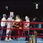Zwei Boxer im Ring, einer trägt amerikanische Flaggenkleidung, der andere einen gelben Umhang. Zuschauer im Hintergrund.