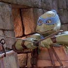 Keine sichtbaren Texte. Eine grüne Schildkröte mit blauer Maske hält zwei Schwerter vor sich, in einer steinernen Umgebung.