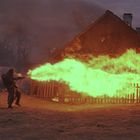 Ein Soldat feuert einen Flammenwerfer auf ein brennendes Holzhaus, große Flammen in lebhaften Farben.