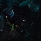 Eine dunkle und regnerische Szene von oben mit einem alten Haus, beleuchteten Fenstern und einem Auto im Hof.