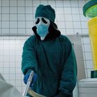 "THE STUFF" steht auf einem gelben Behälter. Person im OP-Kittel mit Maske und gruseligem Gesicht in einem Kachelraum.