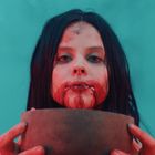 Es gibt keinen Text. Eine Frau mit blutigem Gesicht hält eine Schale vor einem blauen Hintergrund.