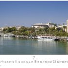 „Sevilla“ steht unten links. Im Hintergrund ist eine Flusspromenade mit Booten und Palmen zu sehen.