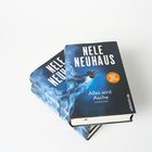 Auf einem Tisch liegen zwei Bücher mit dem Titel "Alles wird Asche" von Nele Neuhaus, dekoriert mit einem Vogel.