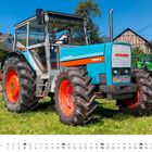 Traktor mit der Aufschrift "EICHER 3075" auf blauer Motorhaube vor grüner Wiese. Unten Kalender: Juli mit Traktor-Ikonen.