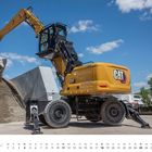 Unten steht "JULI" mit einem Kalender. Ein gelber Bagger mit "CAT"-Logo hebt Kies in einer industriellen Umgebung.