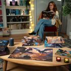 Verschiedene Puzzleschachteln, eine Frau sitzt und liest ein Buch, gemütliches Wohnzimmer mit Bücherregalen und Kerzen.