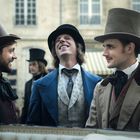 Drei Männer in historischer Kleidung und Zylindern lachen miteinander auf einer Straße vor einem Gebäude mit Fenstern.
