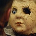 Eine beschädigte Puppenmaske mit blondem Haar und einem kaputten Auge, das bläulich blickt. Das Gesicht zeigt Risse.