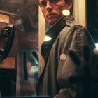 Ein Mann in einem Trenchcoat steht in einer Telefonzelle. Hintergrund unscharf, Lichter und Graffiti erkennbar.
